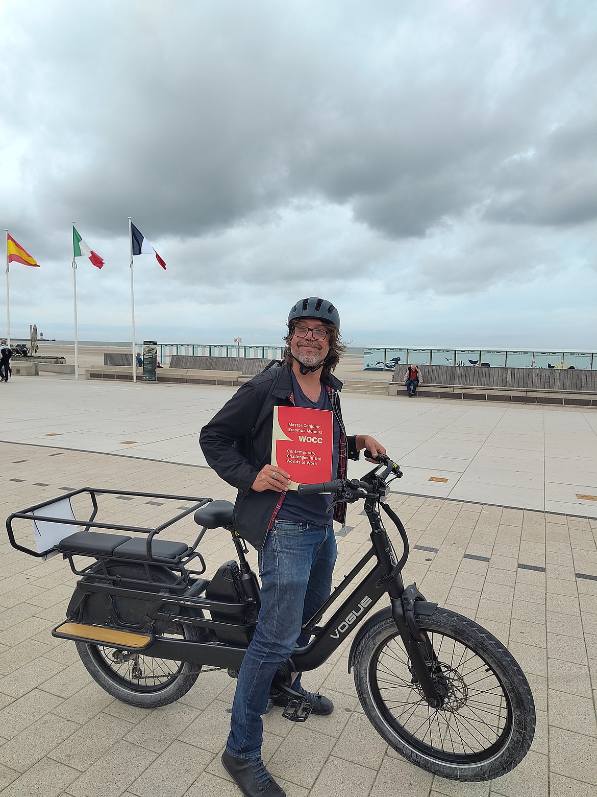 Photo of Director José Calderon Gil on a bike, holding the red and beige WOCC Masters folder, on the seafront in Dunkirk