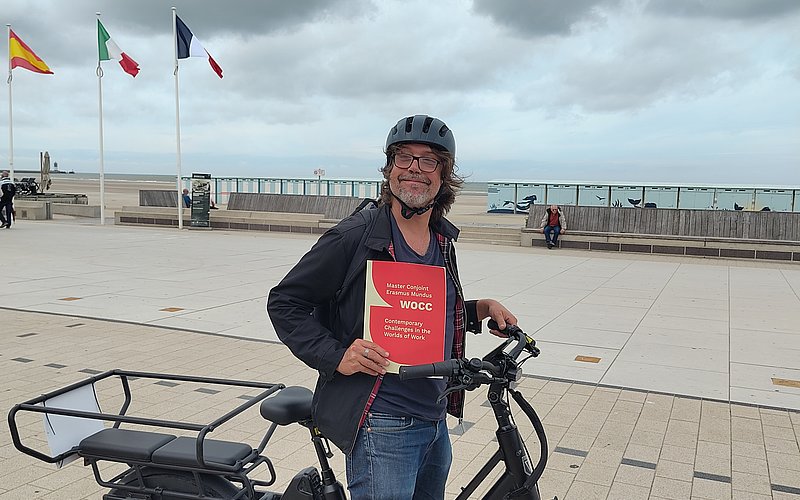 Photo du directeur José Calderon Gil à vélo tenant la pochette rouge et beige du Master WOCC sur le bord de mer de Dunkerque 